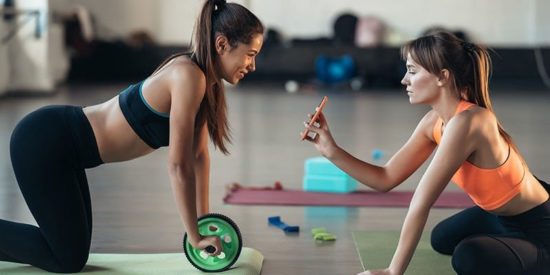 Sporthygiene_Web800-800×400.jpg Zwei Frauen trainieren in einem Fitnessstudio, eine benutzt ein grünes Trainingsgerät und die andere filmt mit ihrem Handy.