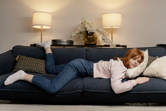 Eine junge Frau mit roten Haaren liegt entspannt auf einem blauen Sofa, umgeben von Kissen und Lampen.