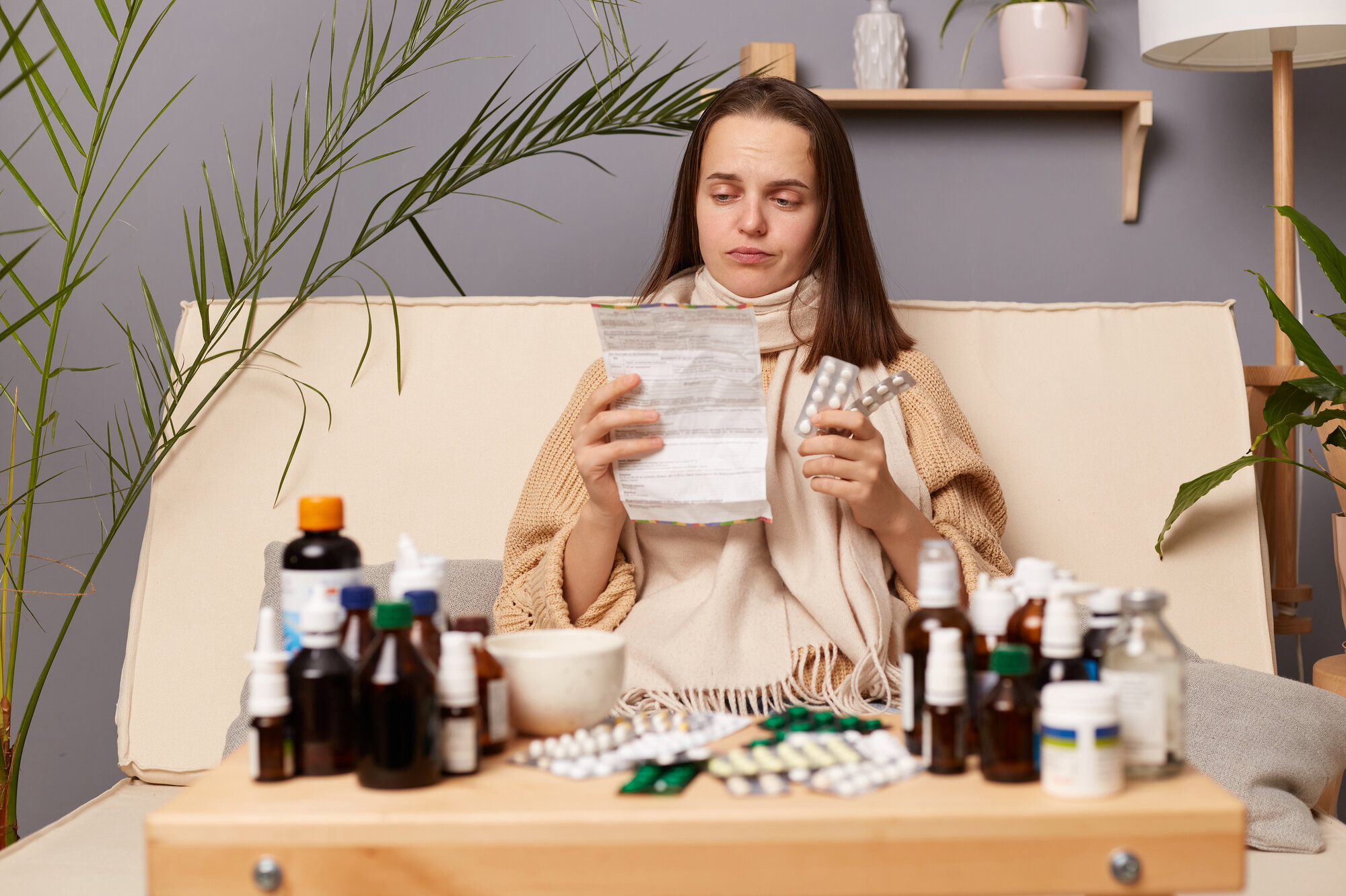 selbstmedikation.jpg Eine kranke Frau sitzt auf einem Sofa, umgeben von verschiedenen Medikamenten und hält einen Zettel sowie eine Medikamentenpackung in der Hand.