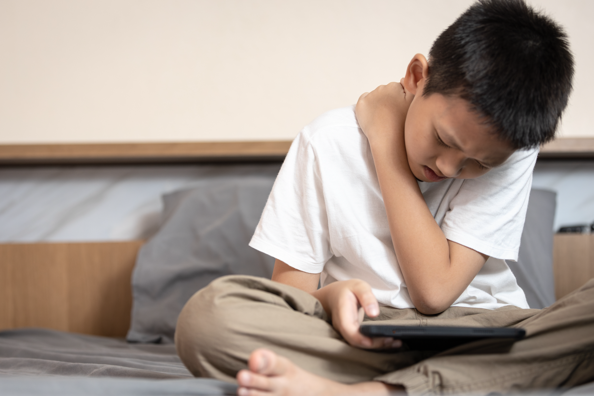 Ein Junge sitzt auf einem Bett, hält ein Tablet und fasst sich schmerzverzerrt an den Hals.