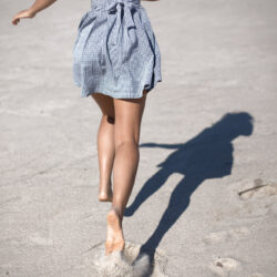Eine Person in einem karierten Kleid läuft barfuß über einen sandigen Strand, während ihr Schatten sichtbar ist.