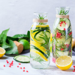 Zwei Glaskaraffen mit Wasser, Gurken, Zitronen und Kräutern, umgeben von frischen Zutaten auf einer Marmorfläche.