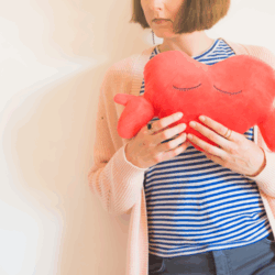 Eine Person hält ein rotes herzförmiges Kissen mit geschlossenen Augen, gekleidet in gestreiftes Oberteil und eine helle Strickjacke.