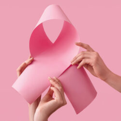 Zwei Hände halten ein rosa Papier, das zu einer Schleife geformt ist, vor einem pinken Hintergrund.
